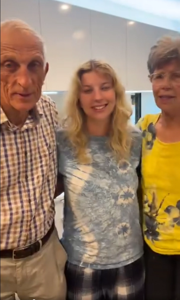 Stephanie’s parents standing with their granddaughter, smiling warmly together in a candid family moment indoors.