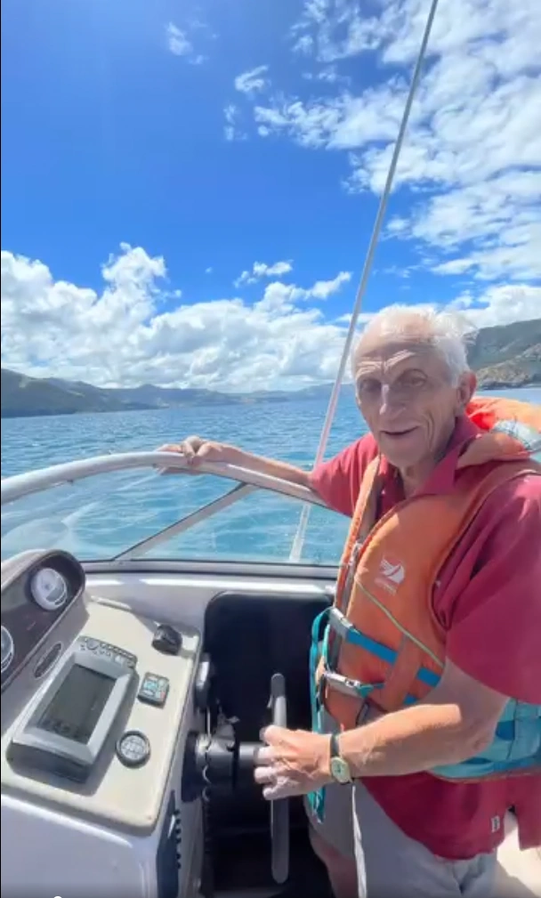 Stephanie’s father Donn smiling while steering a boat, wearing a life jacket, enjoying a bright day on the water.