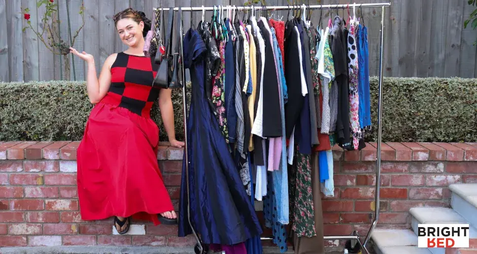 Olivia Hay-Smith smiling in a red and black dress poses beside a full rack of colourful vintage clothing displayed outdoors against a brick wall.