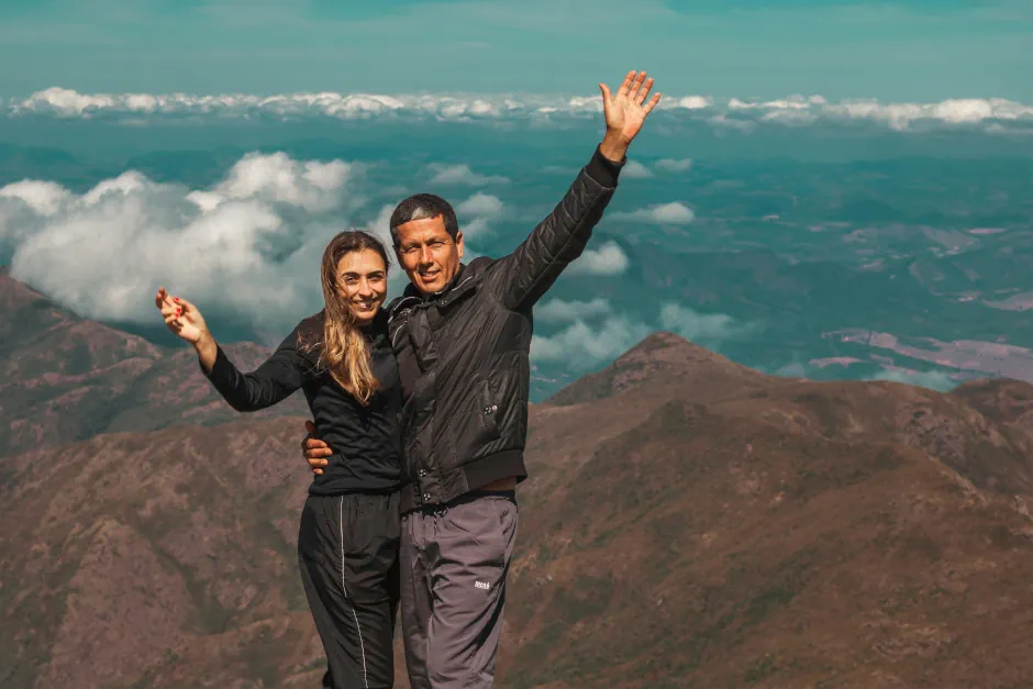 Man and woman celebrating at a mountain summit, dressed in casual outdoor clothing and smiling with arms raised against a scenic landscape.