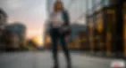 Woman in smart casual winter outfit with blazer, sweater, jeans, and boots, standing confidently in a city at sunset.