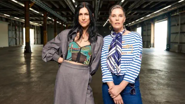 Two women standing confidently in an industrial space, wearing bold, eclectic outfits that showcase a creative and expressive style personality.