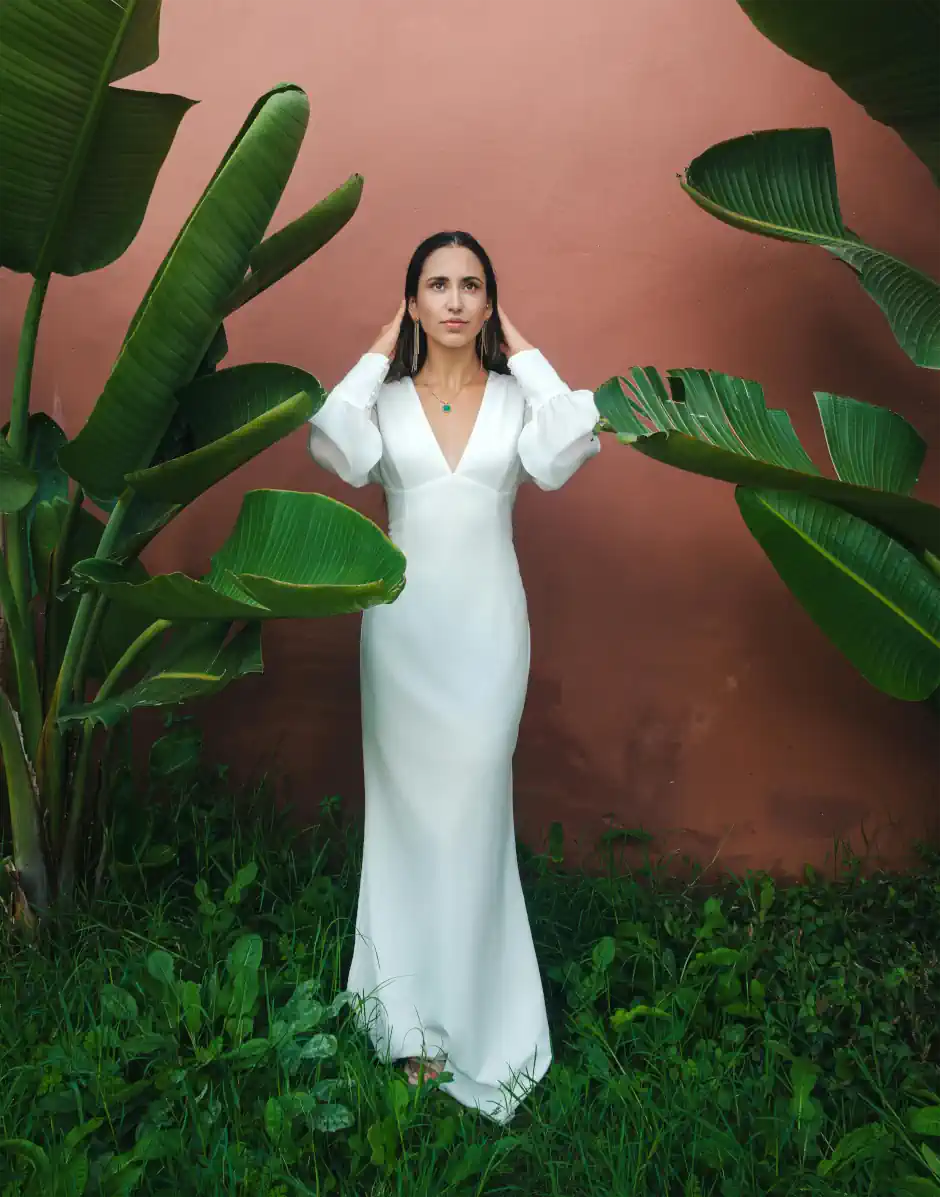 Woman standing between large tropical leaves, wearing a long white dress with puff sleeves, creating a soft, romantic and elegant feminine style.