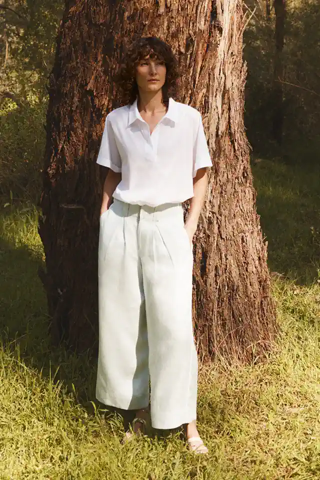 Woman standing against a tree in a relaxed white shirt and wide-leg trousers, showcasing a simple, natural and effortlessly comfortable style.