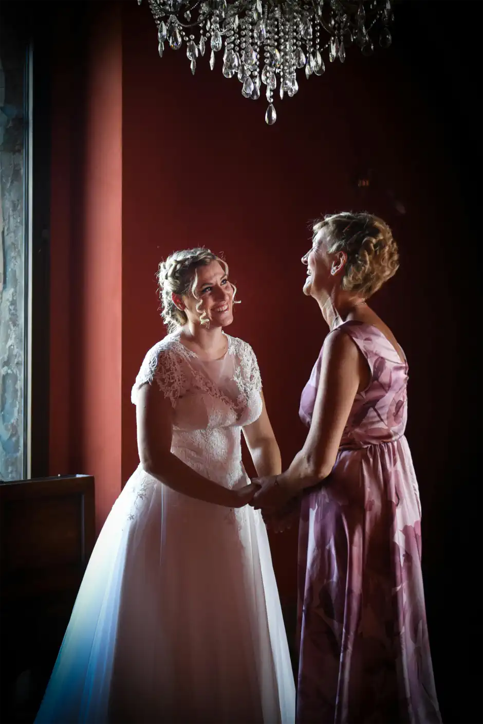 Bride holding hands with an older woman indoors, both smiling in elegant wedding outfits beneath a chandelier.