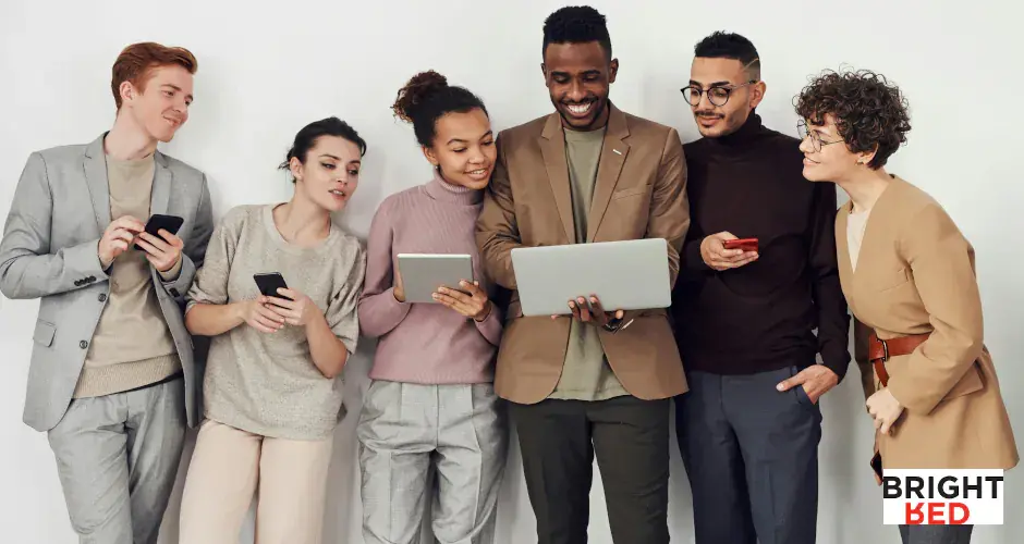 Diverse group of people standing together, smiling and sharing content on phones and a laptop in a relaxed setting.