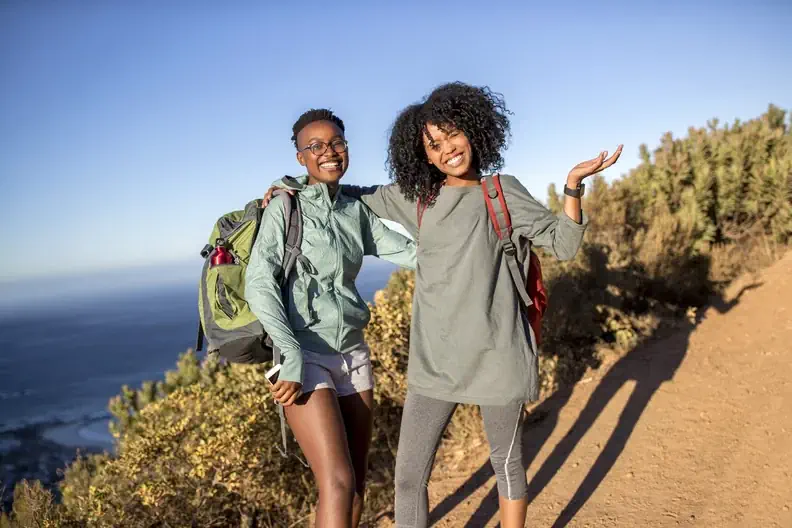 Two women smiling on a sunny hiking trail, wearing relaxed activewear and backpacks, expressing an easy-going, outdoorsy natural style.