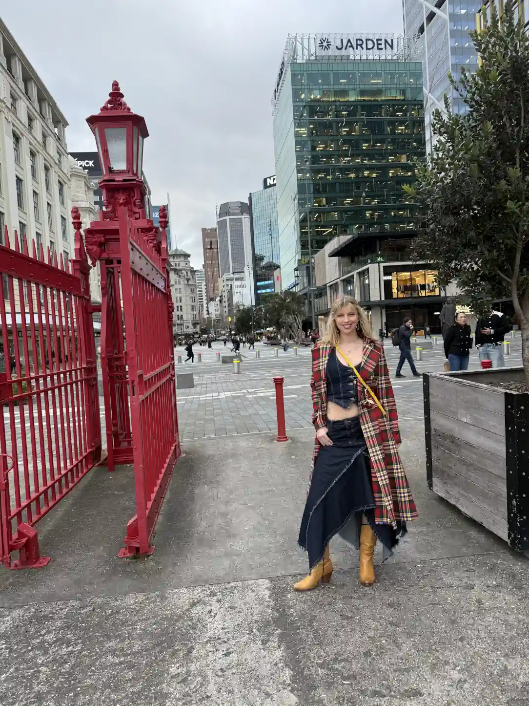 Poppy poses in Auckland wearing a tartan coat, denim outfit, and tan boots, standing near a red gate with city buildings behind.