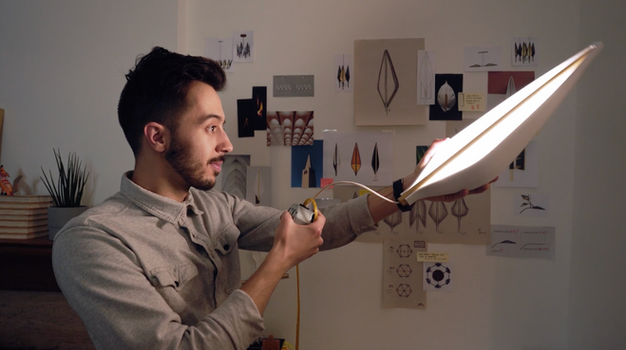A man, Jake Oliveira, holding part of a light to test it. He is standing in front of a wall with various sketches hung up.