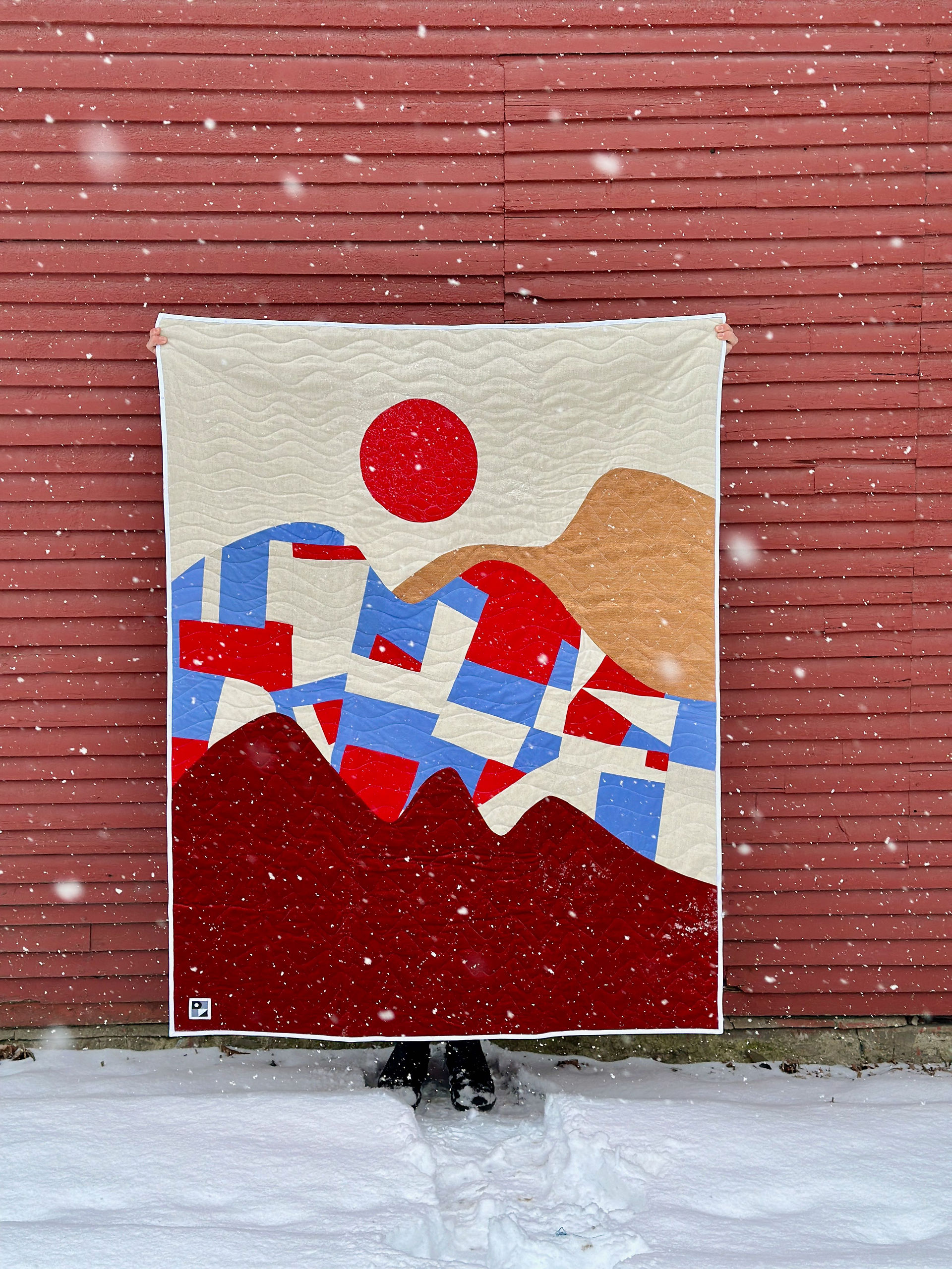 A person holding up a quilt with am abstract landscape of mountains.