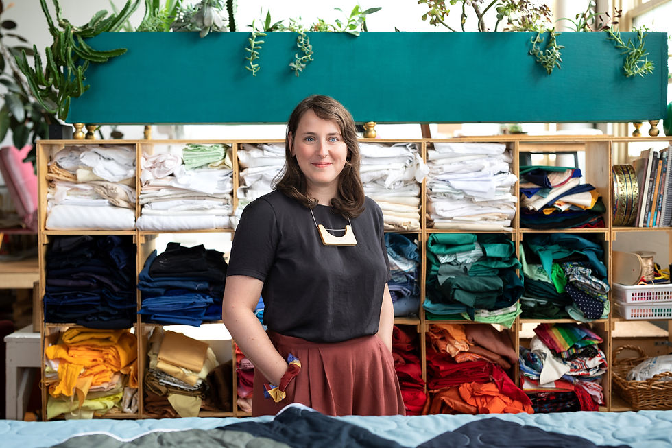 Photo of Marilyn Armand, a woman wearing a black shirt and maroon pants, standing in front of folded fabrics in different colours. 