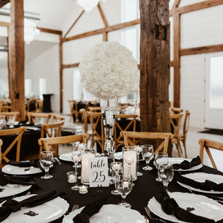 Lush white orchid centerpiece towering over a reception table, set against a background of soft, ambient lighting in the White Barn. The table is dressed in black, complemented by white plates with custom black napkins, creating an elegant and inviting wedding decor.