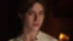 Woman from Mafia: The Old Country with a gentle expression, wearing a floral lace blouse. Dimly lit background with warm candlelight creating a cozy atmosphere.