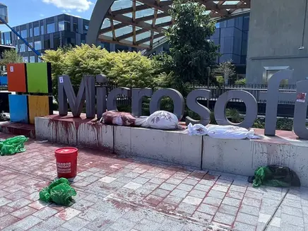 Protest scene at Microsoft sign with red paint, tarp-covered objects, and scattered items. Urban setting with lush greenery.