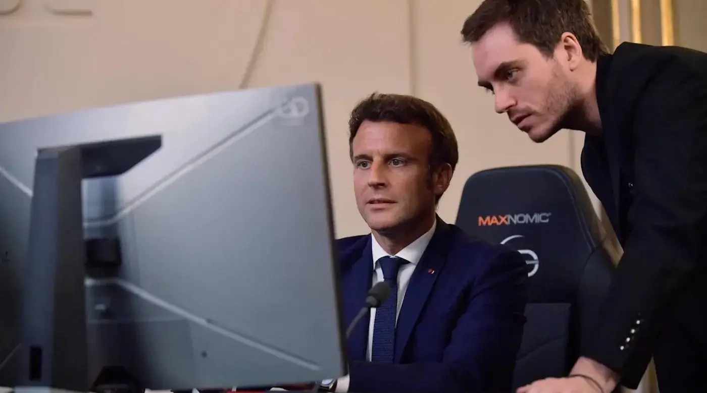 Two men in suits focus on a desktop monitor in an office; one leans over the other. MAXNOMIC and 3D appear on gear.