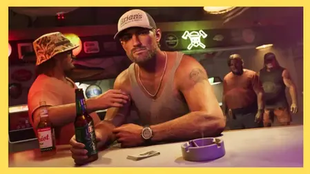 Man in tank top and cap holding beer at a bar, others in background. Neon signs, tattoos, and relaxed atmosphere. Ashtray on counter.