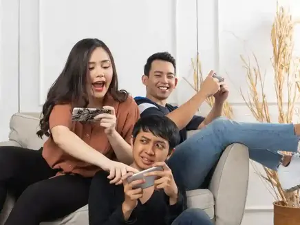 Three people playfully gaming on phones and controllers, sitting on a beige sofa. The setting is bright with dried plants in the background.