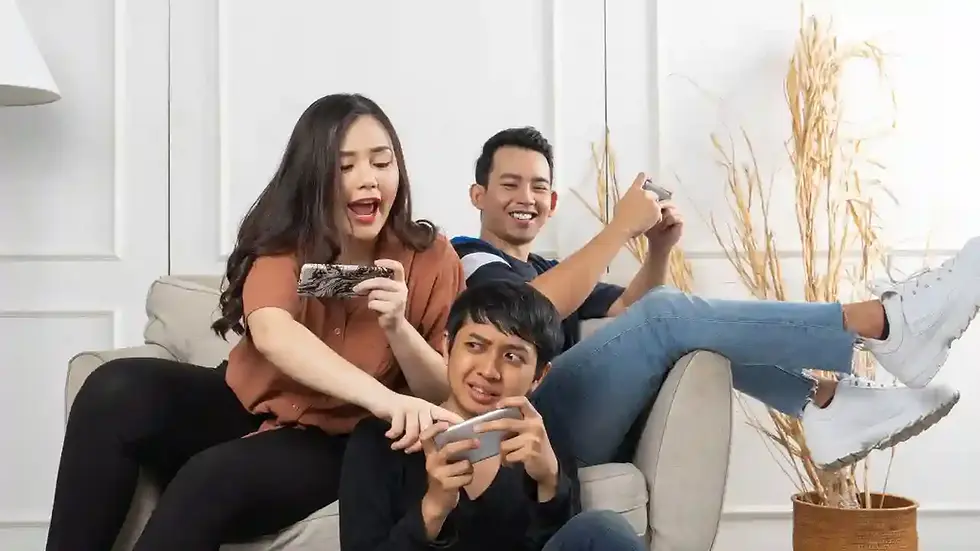 Three people playfully gaming on phones and controllers, sitting on a beige sofa. The setting is bright with dried plants in the background.