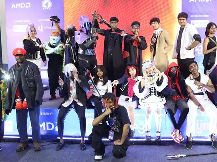 Group of people in colorful costumes posing on stage. Vibrant lights, enthusiastic mood. Background features AMD logos and text. its from Chennai Thiruvizha.