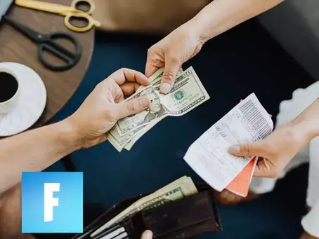 Hands exchanging money and a receipt over a wooden table with scissors, a coffee cup, and a blue square with an "F" on it.