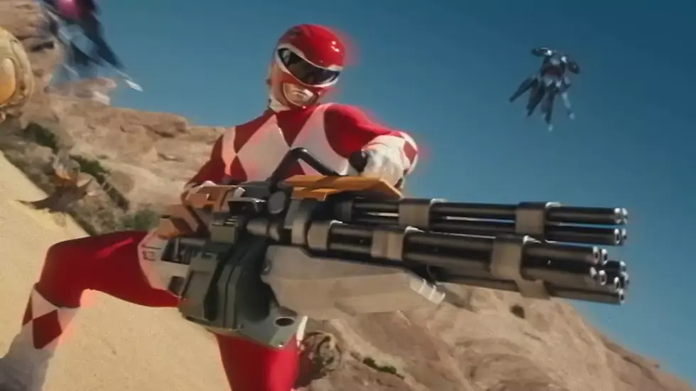 Red superhero in a desert landscape, holding a large futuristic weapon. Blue sky and scattered rocks in the background. Dynamic action pose. its Power Rangers in Fortnite c6s4.