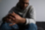 Ein Mann sitze auf einem Sofa, seine Ellenbogen stützt er auf seine Knien ab. Seine Hände berühren sich. Er ist leicht nach vorne gebeugt und schaut nachdenklich auf den Boden. Nur die Hände sind klar erkennbar, der Rest des Bildes ist weichgezeichnet..