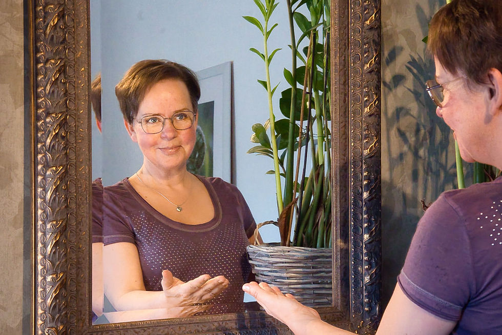 Annette Duveroth stands before an ornately framed mirror. In the reflection, her gaze meets her own. She smiles gently, hands lightly touching the glass. A green plant is visible in the background.