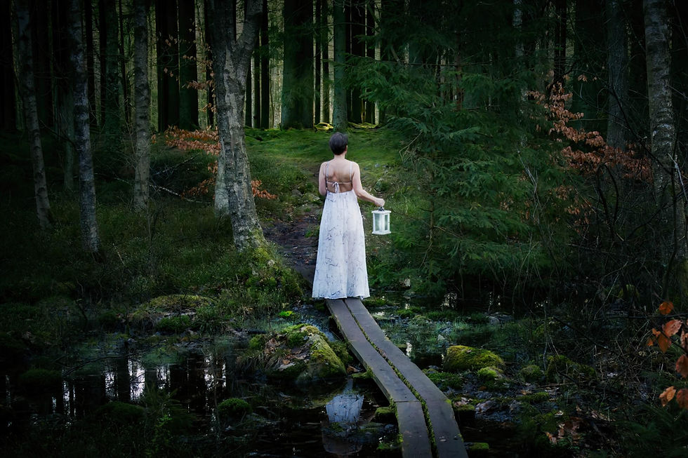 Woman in a white dress walking along a narrow boardwalk through a mossy forest, carrying a lantern. Photographed by Annette Duveroth.