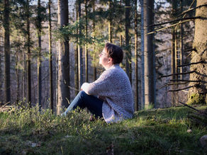 En kvinna sitter på en mossbevuxen sten i skogen och blickar uppåt i mjukt kvällsljus, omgiven av höga granar.