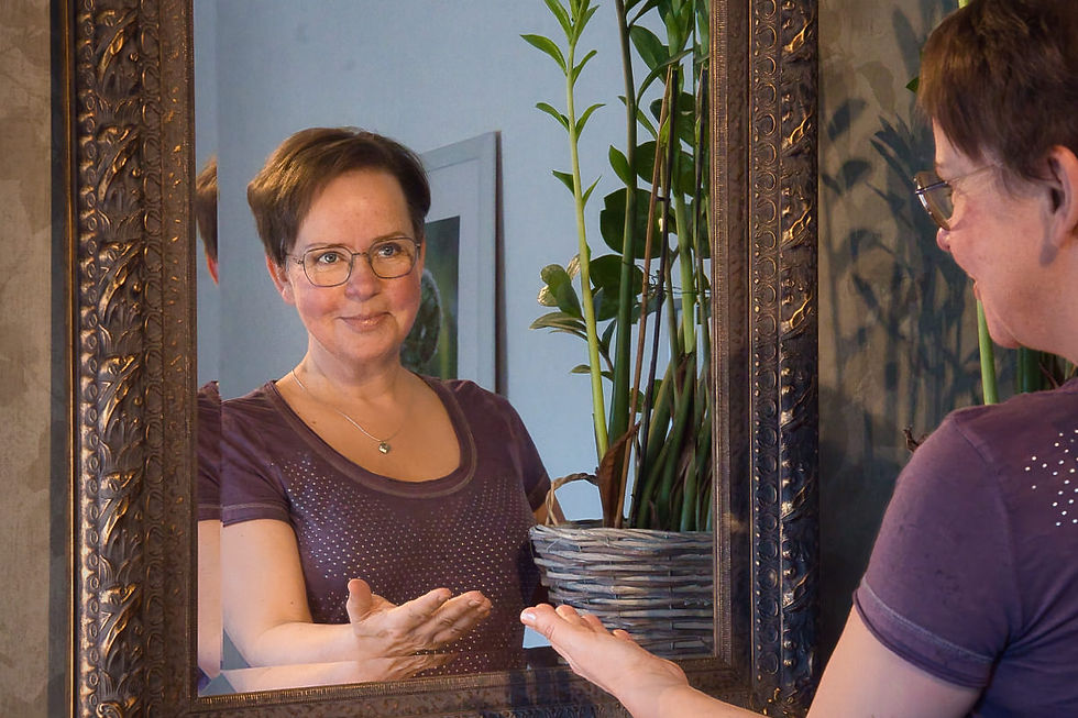 Annette Duveroth stands before an ornately framed mirror. In the reflection, her gaze meets her own. She smiles gently, hands lightly touching the glass. A green plant is visible in the background.