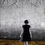 Thumbnail: Woman in black and white walking trough withered grass with shoes in her hand and branches above her, towards dark grey city.
