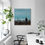 Thumbnail: Square painting with boy walking through field of faded dark brown flowers under blue sky, on white wall next to window