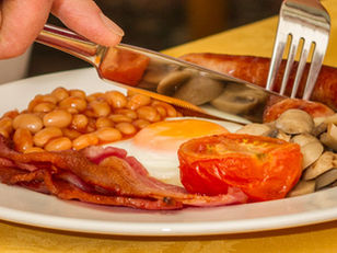 The Claremont St Annes Breakfast: Plate with bacon, eggs, beans, mushrooms, sausage, and tomato on a yellow tablecloth. A hand cuts with a knife and fork.