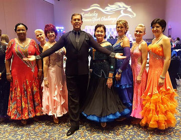 Group of dancers with instructor in costumes smiling during a ballroom dance competition.