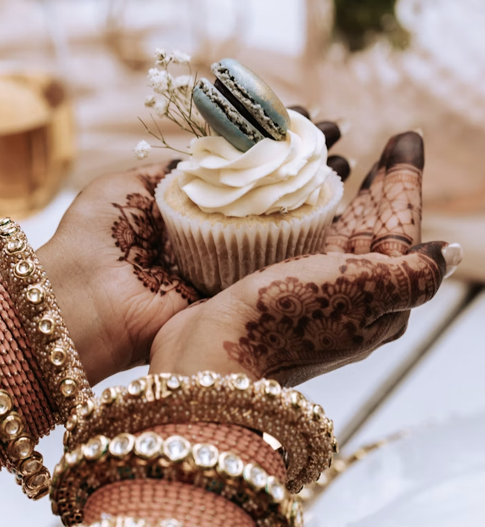 Close-up of hands holding a cupcake topped with a macaron, featuring delicate front hand mehndi designs and glittering bangles.