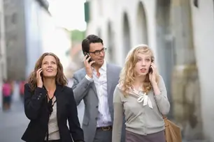 Three people walking on a street, each talking on a phone. They appear engaged, with a blurred urban background and arches.