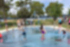 Kids play joyfully in a water park with sprinklers. Adults watch nearby. Green trees and a lake are in the background under a blue sky.