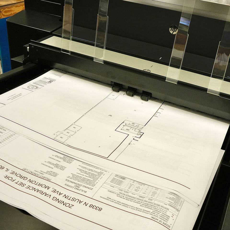 A black-and-white CAD architectural drawing is printed on a large-format plotter for construction and project documentation.