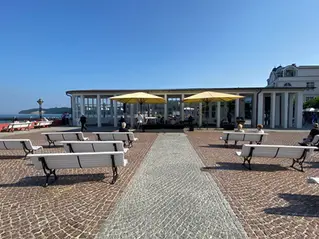 Zwei gelbe Großschirme auf einem gepflasterten Platz vor einem Pavillon, davor weiße Bänke