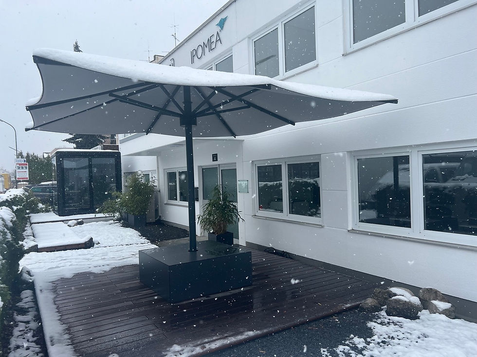Großschirm mit Schnee auf dem Dach, auf einem dunklen Sitzelement vor einem weißen Firmengebäude bei Schneefall