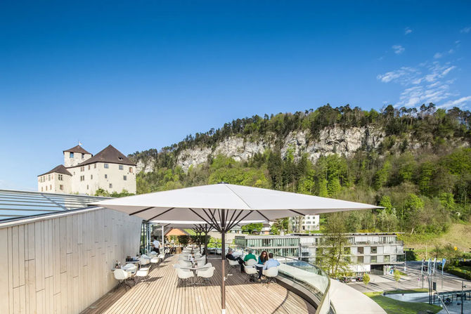 Sehr großer quadratischer Sonnenschirm mit hellem Dach und einem Mast über mehreren Tischgruppen auf einer Dachterrasse