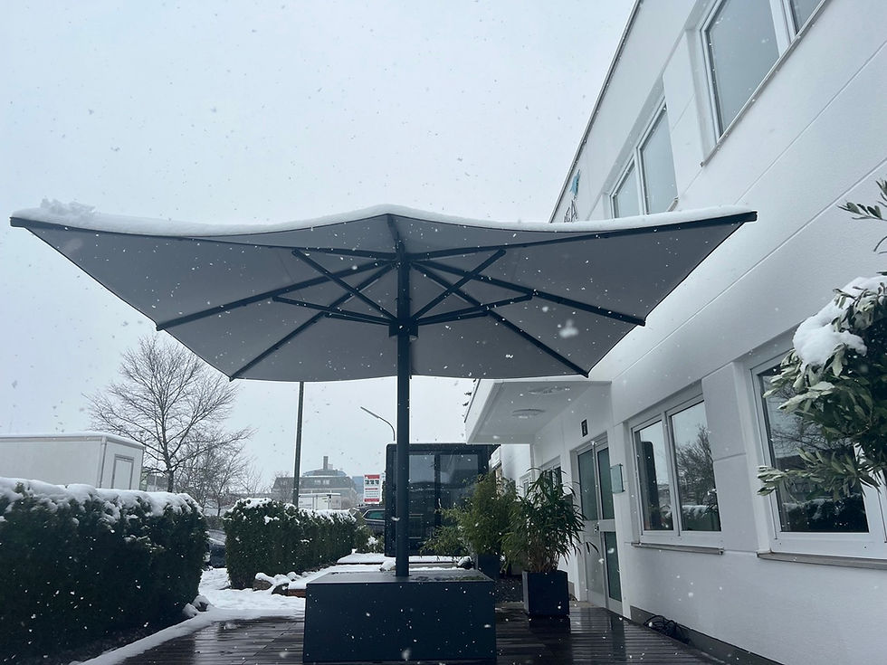 Geöffneter Großschirm mit Schnee auf dem Dach vor einem weißen Firmengebäude, von unten bei Schneefall fotografiert