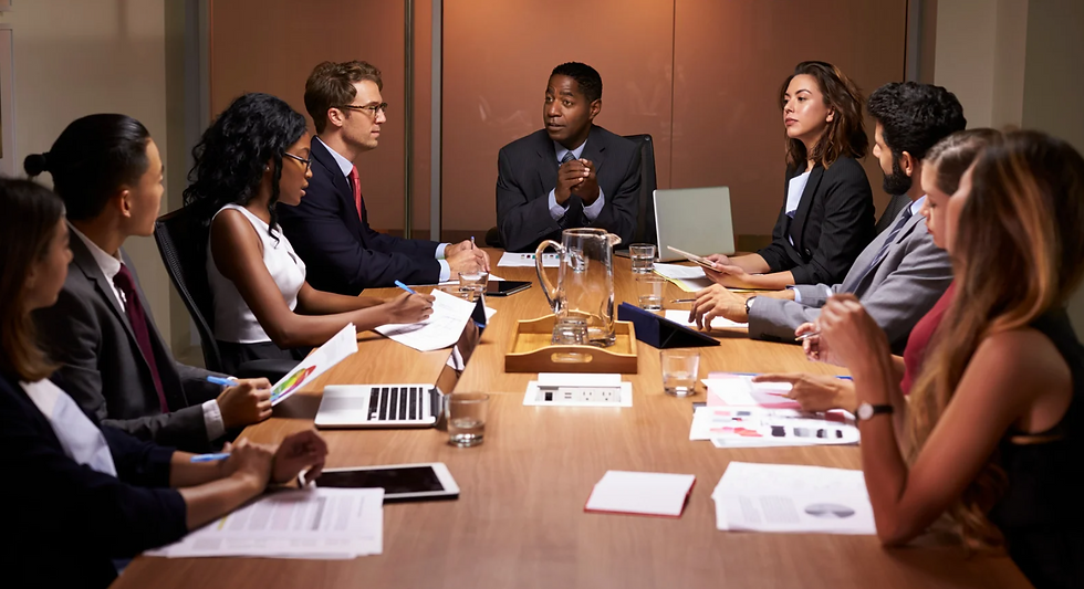 A group of people in business attire sit around a conference table, engaged in discussion. Papers and laptops are scattered on the table.