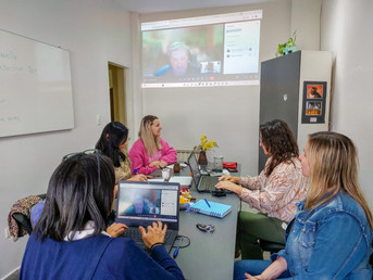 Foto. Mesa de trabajo en ambiente cerrado e iluminado. Cinco mujeres alrededor de la mesa miran hacia el frente de la sala donde hay una pantalla y en ella se ve a otra persona que está conectada virtualmente en la reunión.