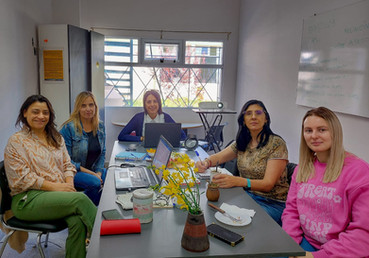 Foto. Mesa de trabajo en ambiente cerrado e iluminado. Cinco mujeres alrededor de la mesa miran hacia la cámara.