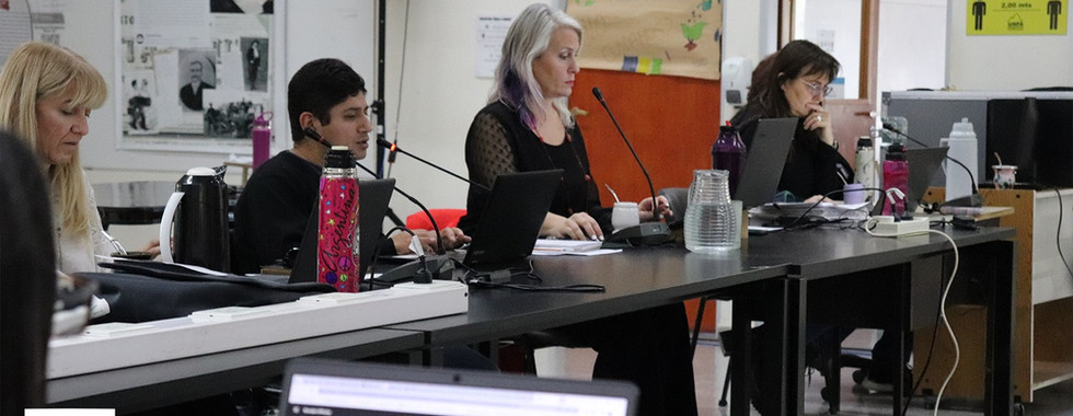 Reunión de Consejo de Unidad.  Grupo de personas sentadas detrás de una mesa alargada, cada una con un micrófono delante y una notebook.