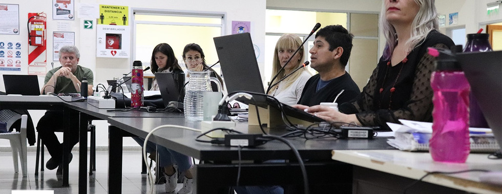 Reunión de Consejo de Unidad.  Grupo de personas sentadas detrás de una mesa alargada, cada una con un micrófono delante y una notebook.