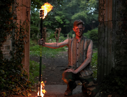 Man crouched down in the woods holding staff on fire on both ends looking directly into camera