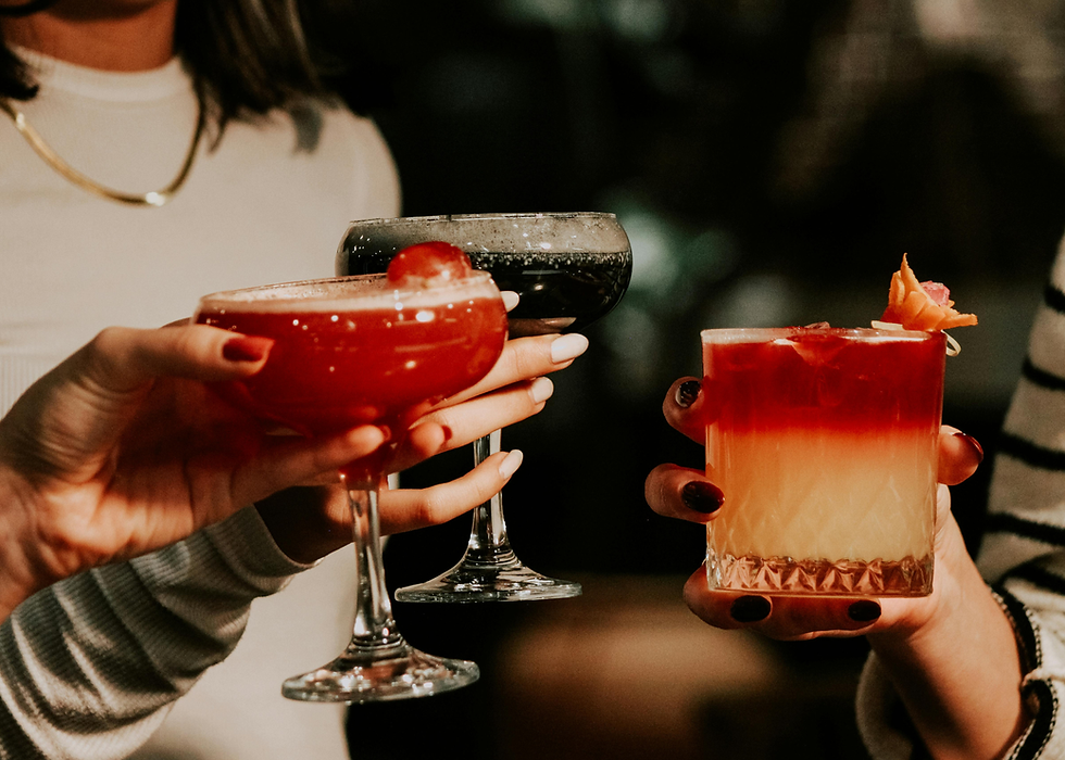 Three hands holding colourful cocktails in various glasses, set against a blurred background. The mood is celebratory and warm.