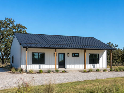 Small white barndominium guest house with black metal roof, front porch, gravel drive, and native landscaping.